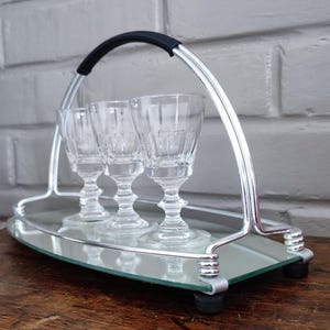 May include: A silver-toned serving tray with a black handle holds three clear, stemmed glasses. The tray has a mirrored surface and is set on a wooden table. Additional glasses are visible to the left.