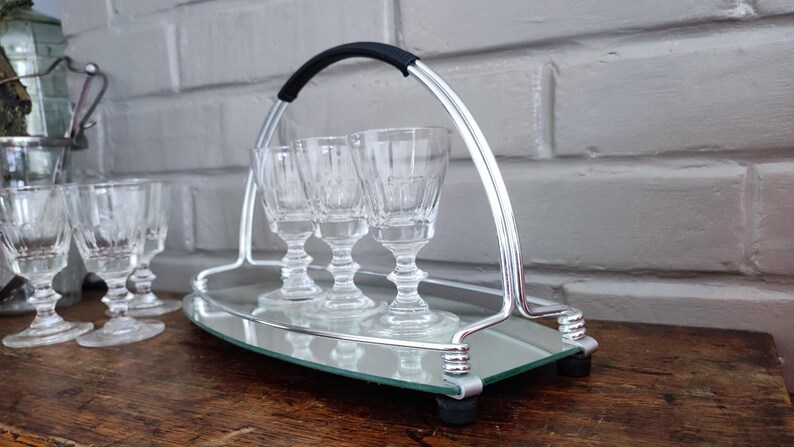 May include: A silver-toned serving tray with a black handle holds three clear, stemmed glasses. The tray has a mirrored surface and is set on a wooden table. Additional glasses are visible to the left.