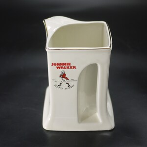 May include: A white ceramic Johnnie Walker Scotch Whisky pitcher with a gold rim. The pitcher features the Johnnie Walker logo in red and black, with the text "Johnnie Walker Scotch Whisky".