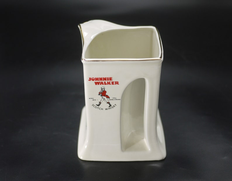May include: A white ceramic Johnnie Walker Scotch Whisky pitcher with a gold rim. The pitcher features the Johnnie Walker logo in red and black, with the text "Johnnie Walker Scotch Whisky".
