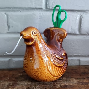 Könnte beinhalten: Ein glänzender, brauner Keramik-Garnhalter in Form eines Huhns mit strukturierter Oberfläche. Ein weißer Faden kommt aus dem Schnabel des Huhns und eine leuchtend grüne Schere steckt in einem Schlitz auf dem Rücken der Keramikvogel-Figur.