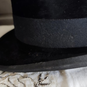 May include: A black top hat featuring a textured velvet-like finish on the crown and brim. The hat includes a wide, matte black grosgrain ribbon band encircling the base of the crown and a matching black fabric trim along the edge of the curved brim.