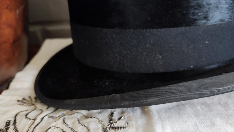 May include: A black top hat featuring a textured velvet-like finish on the crown and brim. The hat includes a wide, matte black grosgrain ribbon band encircling the base of the crown and a matching black fabric trim along the edge of the curved brim.