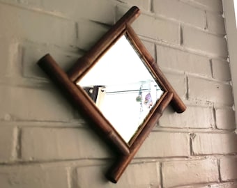 May include: A square mirror with a dark brown bamboo frame, positioned at a diamond angle. The bamboo stalks extend beyond the corners of the mirror frame, creating a cross-like structure. The reflective surface shows a clear indoor reflection.