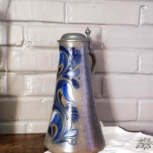 Peut inclure: Une grande chope à bière conique en céramique avec un vernis beige moucheté et un couvercle en étain métallique. L'avant présente un motif floral en relief peint à la main dans des tons bleu foncé, bleu clair et marron, avec une bordure en zigzag.