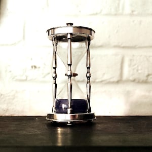 May include: An hourglass featuring a polished silver-toned metal frame with four slender vertical supports and circular top and bottom plates. The clear glass bulb contains dark blue sand, currently settled in the bottom chamber.