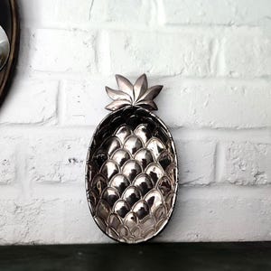 Op de afbeelding: Een zilverkleurige decoratieve schaal in de vorm van een ananas. De schaal heeft een gedetailleerd, gestructureerd oppervlak dat de schil van een ananas nabootst. De schaal is ovaal van vorm met een bladachtige bovenkant.