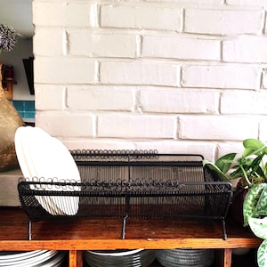 May include: A black wire dish drying rack with a rectangular frame and decorative coiled wire edges. The rack features a grid base and vertical wire supports, currently holding several white plates on the left side, supported by small metal legs.