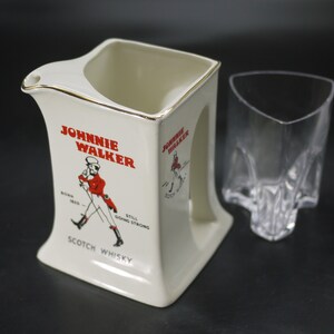 May include: Cream-colored ceramic pitcher and a clear glass. The pitcher has a gold rim and features the "Johnnie Walker" logo in red, with an illustration of a man in a red coat. The glass has a unique, angular design.