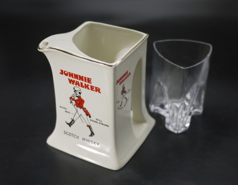 May include: Cream-colored ceramic pitcher and a clear glass. The pitcher has a gold rim and features the "Johnnie Walker" logo in red, with an illustration of a man in a red coat. The glass has a unique, angular design.