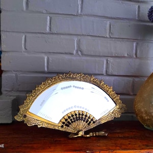 Peut inclure: Miroir décoratif en forme d'éventail avec un cadre doré orné. Le miroir a une surface blanche avec un motif de petits points et de lignes. L'éventail repose sur une surface en bois devant un mur de briques blanches.