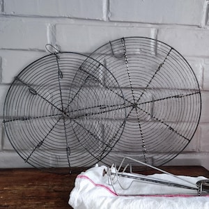 May include: Two round, black wire cooling racks with a radial design, resting on a wooden surface. A white cloth with a red trim and a whisk are in the foreground. The background features a white brick wall.