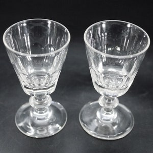 May include: Two clear, stemmed glasses with a faceted bowl design. The glasses have a round base and a decorative stem with a bulbous section. The glasses are likely for serving wine or other beverages.