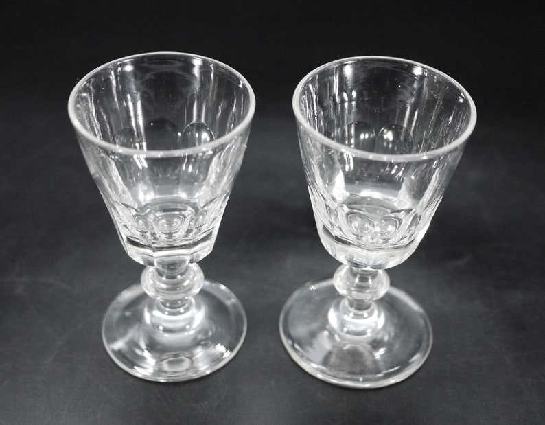 May include: Two clear, stemmed glasses with a faceted bowl design. The glasses have a round base and a decorative stem with a bulbous section. The glasses are likely for serving wine or other beverages.