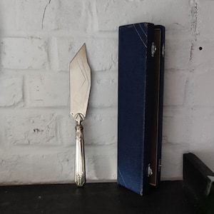 May include: A silver cake server with a decorative handle and a geometric blade design. Beside it is a tall, dark blue box with silver accents, likely for storage. The server is displayed against a white brick wall.