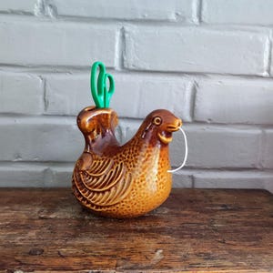 Peut inclure: Un dévidoir à ficelle en céramique émaillée marron en forme de poule avec un motif de plumes texturé. L'objet présente une ouverture sur le dessus contenant une paire de ciseaux verts et un trou au niveau du bec d'où sort une ficelle blanche.