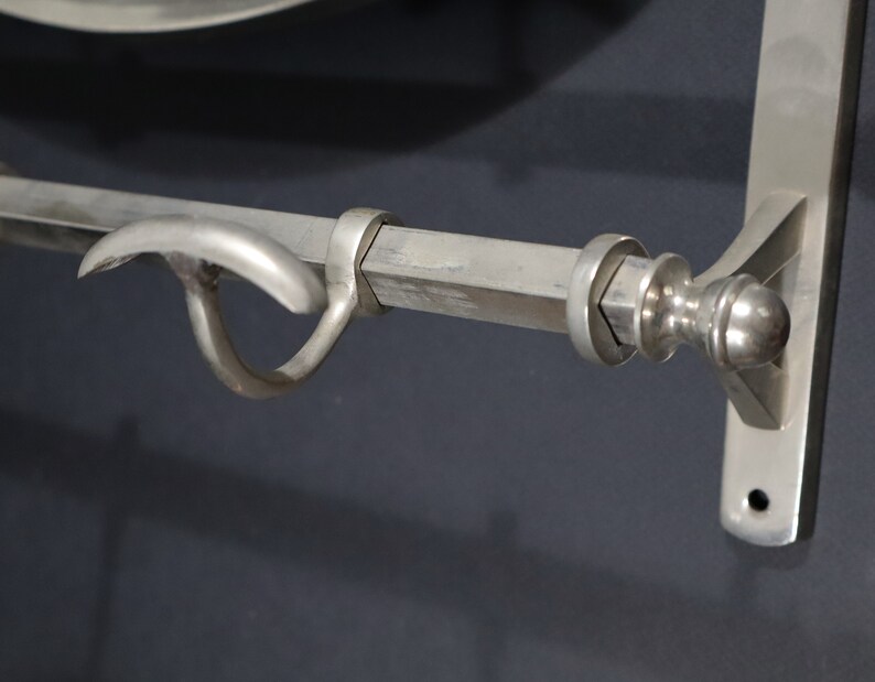 May include: A silver metal towel bar with a hook on the left side and a decorative finial on the right side. The bar is mounted to a wall.