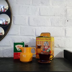 Puede incluir: Una variedad de artículos de café vintage. Una cafetera de cerámica de color amarillo mostaza con tapa marrón oscuro se encuentra junto a una caja de filtro Melitta verde y blanca. También hay un juego de filtro de café marrón y una caja con una ilustración vintage.