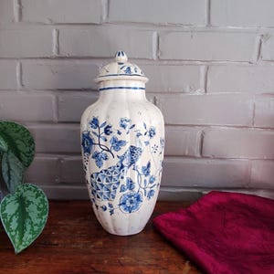 Peut inclure: Un pot à gingembre en céramique couleur crème avec un couvercle amovible, présentant une texture nervurée verticale et un motif peint en bleu représentant un paon entouré de fleurs et de vignes. Le pot a une finition brillante.