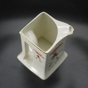 May include: Cream-colored ceramic pitcher with a square base and a handle. The pitcher has a gold trim around the rim and a spout. Red text and a graphic of a person are printed on the side. The pitcher is likely vintage.