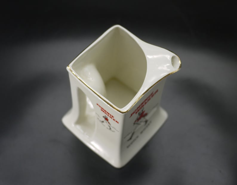 May include: Cream-colored ceramic pitcher with a square base and a handle. The pitcher has a gold trim around the rim and a spout. Red text and a graphic of a person are printed on the side. The pitcher is likely vintage.