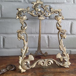 Peut inclure: Un cadre photo en laiton orné présentant des volutes complexes et des motifs de feuilles le long de la bordure. Le cadre possède une ouverture rectangulaire verticale et comprend un support métallique robuste fixé à l'arrière pour une présentation.