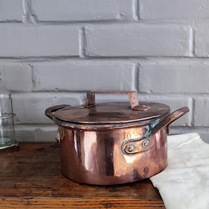 Puede incluir: Una olla de cobre con tapa y dos asas laterales. La olla es redonda y tiene una superficie brillante y reflectante. La tapa tiene un asa en la parte superior. La olla está sobre una superficie de madera.