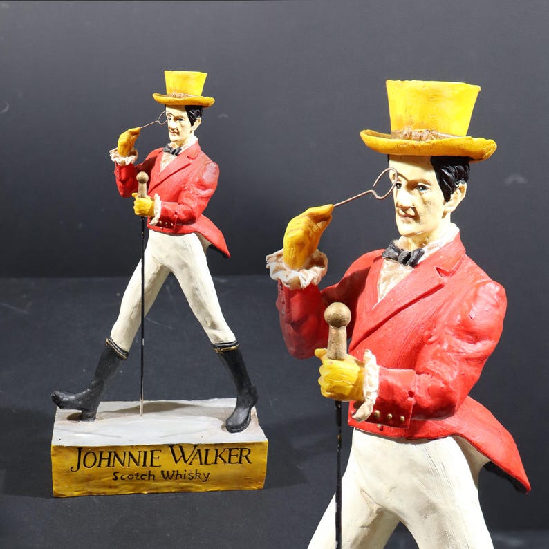 May include: Two Johnnie Walker Scotch Whisky figurines. Each figure is of a man in a red jacket, yellow top hat, and white trousers, holding a cane and monocle. The base of the figurine has the brand name and product description.