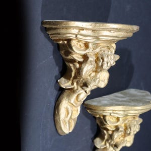 May include: Two gold-painted wall shelves with a decorative, sculpted design resembling a grotesque face. The shelves are made of a lightweight material and have a distressed finish.