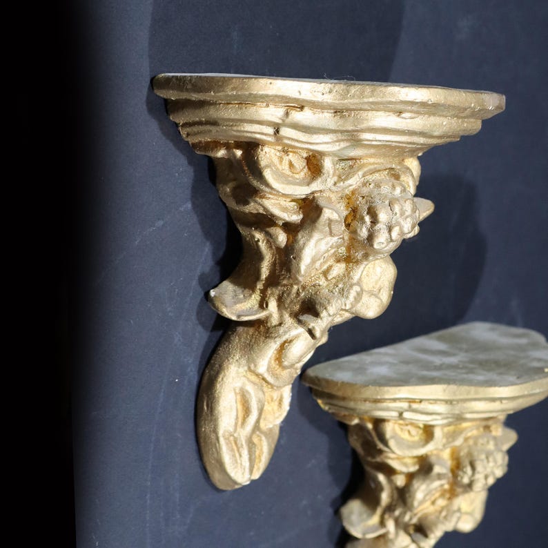 May include: Two gold-painted wall shelves with a decorative, sculpted design resembling a grotesque face. The shelves are made of a lightweight material and have a distressed finish.