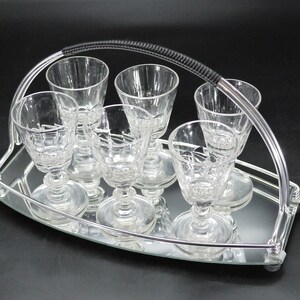May include: A set of six clear crystal cordial glasses on a mirrored tray with a silver metal handle. The glasses have a detailed pattern and are arranged neatly on the tray, perfect for serving drinks.