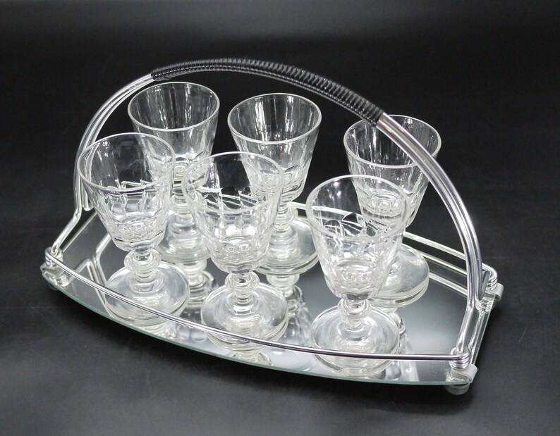 May include: A set of six clear crystal cordial glasses on a mirrored tray with a silver metal handle. The glasses have a detailed pattern and are arranged neatly on the tray, perfect for serving drinks.