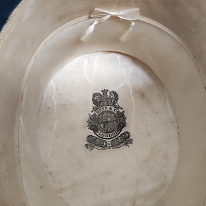May include: The interior of a dark-colored hat featuring a cream-colored lining with a small ribbon bow at the top. A black printed crest logo in the center reads KRESS & CO LONDON. Handwritten text is visible on the lower right side of the lining.