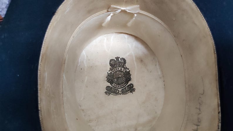May include: The interior of a dark-colored hat featuring a cream-colored lining with a small ribbon bow at the top. A black printed crest logo in the center reads KRESS & CO LONDON. Handwritten text is visible on the lower right side of the lining.