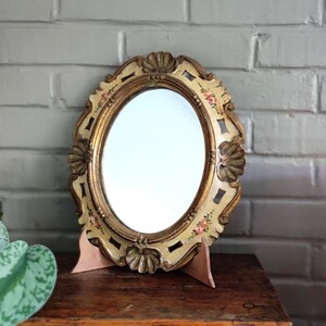 Peut inclure: Miroir de coiffeuse ovale avec un cadre doré et des motifs floraux. Le miroir est soutenu par un support en bois. Le cadre a une base crème avec des bordures dorées et des éléments décoratifs en forme de coquillage.