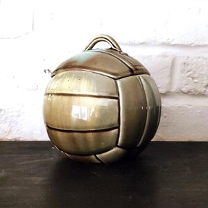 Peut inclure: Un pot en céramique sphérique en forme de ballon de volley-ball avec un vernis brillant aux tons beige et marron. Le couvercle présente une petite poignée arquée sur le dessus, et la surface est segmentée par des lignes en creux.