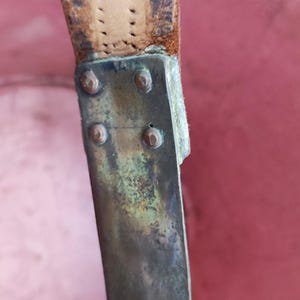 May include: A weathered brown leather strap attached to a tarnished brass metal plate. The rectangular metal plate features four small circular rivets securing it to the worn leather, showing signs of oxidation and age on the metal surface.