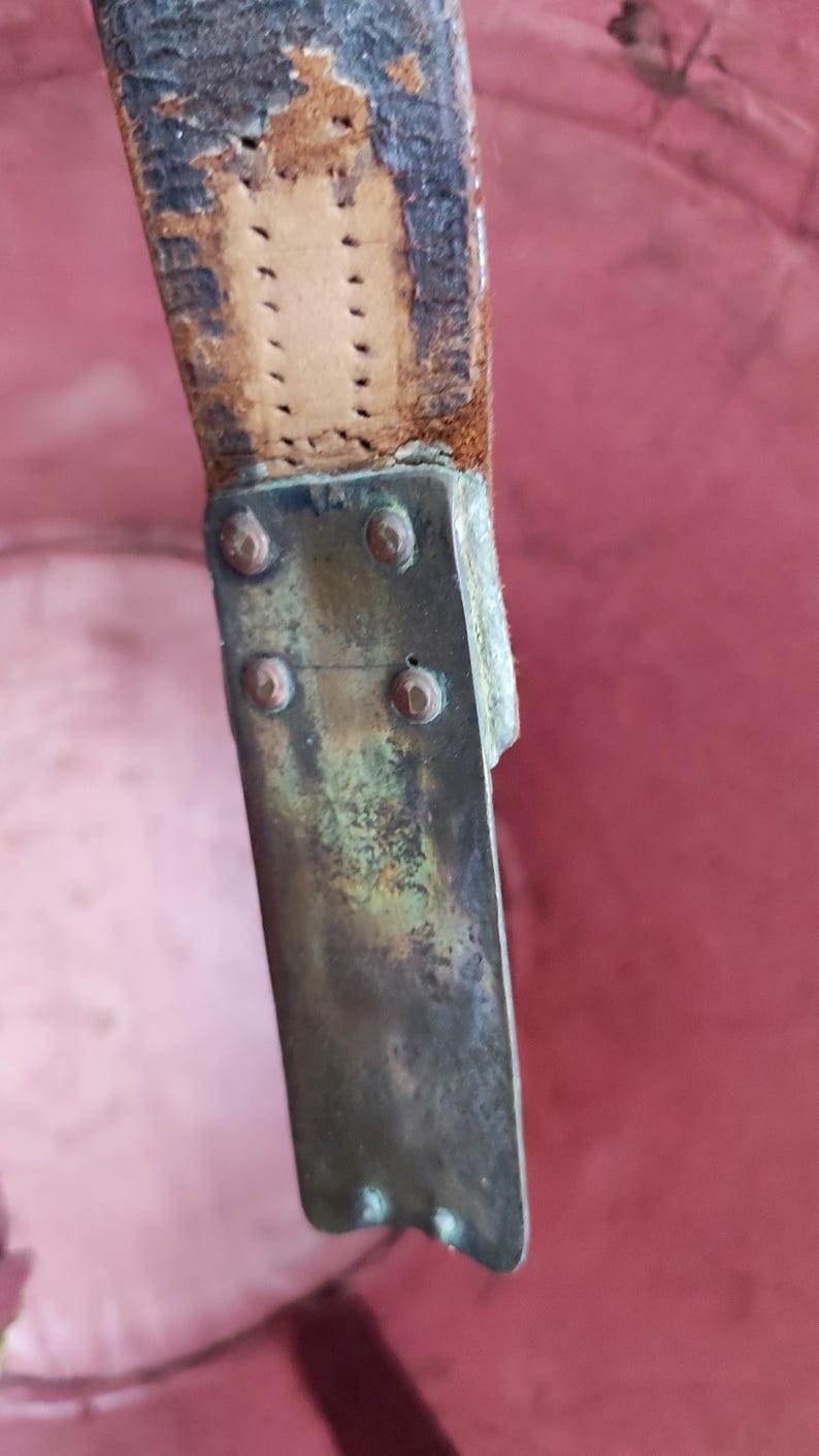 May include: A weathered brown leather strap attached to a tarnished brass metal plate. The rectangular metal plate features four small circular rivets securing it to the worn leather, showing signs of oxidation and age on the metal surface.
