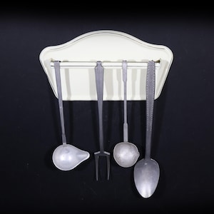 Peut inclure: Un support à ustensiles de cuisine crème avec un dessus décoratif et une bordure noire, contenant cinq ustensiles de cuisine argentés. Les outils comprennent une louche, une fourchette et des cuillères, le tout sur fond noir.