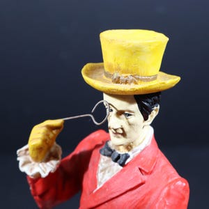 May include: A figurine of a man in a red suit, black bow tie, and yellow top hat, holding a monocle. The figurine has a painted face and black hair. The hat has a brown band.