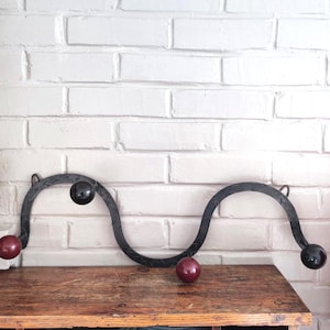 Peut inclure: Un porte-manteau mural décoratif en fer forgé. Le porte-manteau est noir avec un design ondulé et comporte quatre accents sphériques noirs et bordeaux. Le porte-manteau est monté sur un mur de briques blanches.