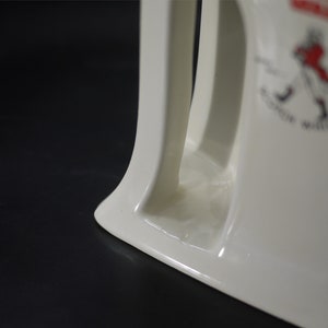 May include: Cream-colored ceramic pitcher with a curved handle and a decorative emblem. The emblem features a figure in red and black, with the words "Scotch Whisky" and other text. The pitcher is on a black surface.