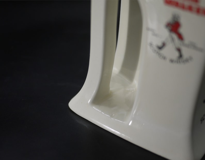 May include: Cream-colored ceramic pitcher with a curved handle and a decorative emblem. The emblem features a figure in red and black, with the words "Scotch Whisky" and other text. The pitcher is on a black surface.