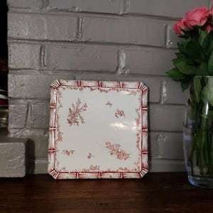 Peut inclure: Assiette carrée en céramique blanche avec un motif floral rouge et une bordure rouge de style bambou. L'assiette est posée sur une surface en bois foncé, avec un mur de briques grises en arrière-plan. Un vase de roses roses est visible à droite.