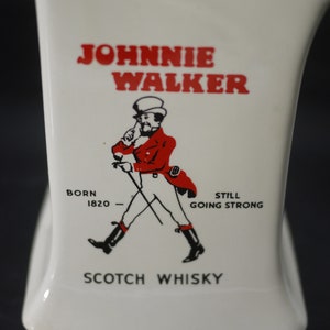 May include: White ceramic bottle with the "Johnnie Walker" logo in red. The logo features a man in a red coat and black boots, walking with a cane. The text "Born 1820" and "Scotch Whisky" are also visible.