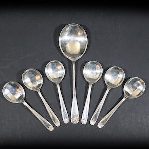 May include: A collection of seven silver spoons, including a large serving spoon and six smaller spoons. The spoons are arranged on a dark surface, reflecting light. The serving spoon is approximately 10 inches long, while the smaller spoons are about 6 inches.