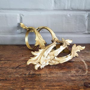 Peut inclure: Deux ronds de serviette en métal doré présentant un motif ornemental en relief de feuilles d'acanthe. Les anneaux possèdent une base circulaire et une extension décorative en forme de feuille qui s'incurve vers l'extérieur de la structure.