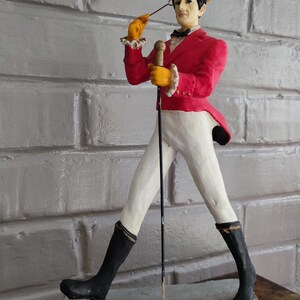 May include: A Johnnie Walker Striding Man figurine, wearing a red jacket, white trousers, and black boots. The figure holds a cane and a yellow top hat. The base reads "JOHNNIE WALKER Scotch Whisky."