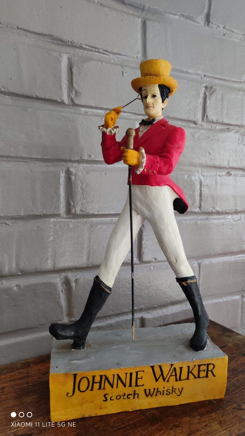 May include: A Johnnie Walker Striding Man figurine, wearing a red jacket, white trousers, and black boots. The figure holds a cane and a yellow top hat. The base reads "JOHNNIE WALKER Scotch Whisky."