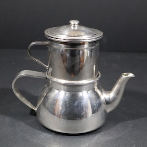 May include: A silver stainless steel coffee pot with a filter and a lid. The pot has a rounded body and a spout. It has two handles.